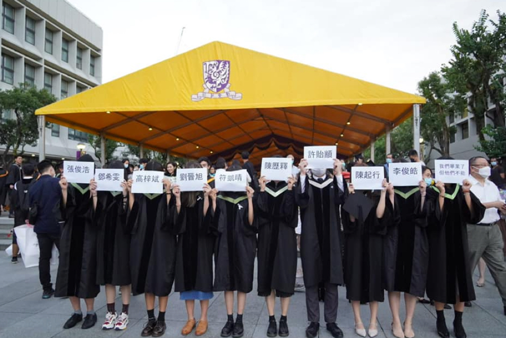 10名中大畢業生傍晚舉起紙牌，寫有9名因2019年中大二號橋衝突而被捕的學生名字，以及「我們畢業了，但他們不能」的字句。（中大學生報Facebook圖片）