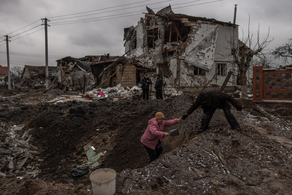 俄羅斯向烏克蘭全境發射導彈，基輔關鍵基礎設施成為襲擊目標。圖為2023年1月26日，在烏克蘭基輔郊外的一個城市型定居點，一名男子幫助一名婦女從受損住宅建築旁邊的一個彈坑中逃生。（Roman Pilipey/Getty Images）