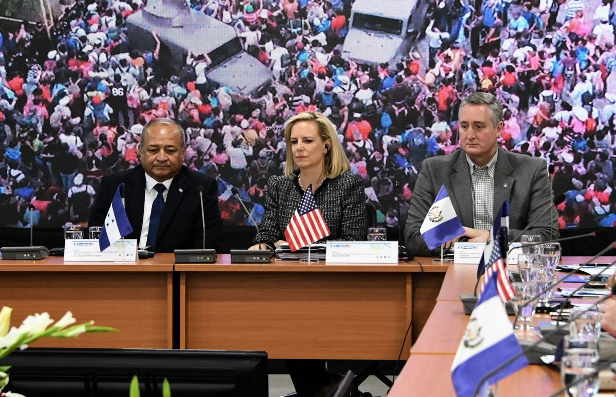 2019年3月27日，美國國土安全部長克爾斯滕・尼爾森（Kirstjen Nielsen）（中） 與洪都拉斯安全部長朱利安・帕切科（Julian Pacheco）（左）和危地馬拉政府部長恩里克‧德根哈特（Enrique Degenhart）（右）出席在洪都拉斯首都特古西加爾巴舉行的「北方三角」（Northern Triangle，薩爾瓦多、危地馬拉和洪都拉斯三個中美洲國家）和美國安全部長會議。屏幕上顯示著一隊中美洲移民的圖片。（ORLANDO SIERRA／AFP／Getty Images）